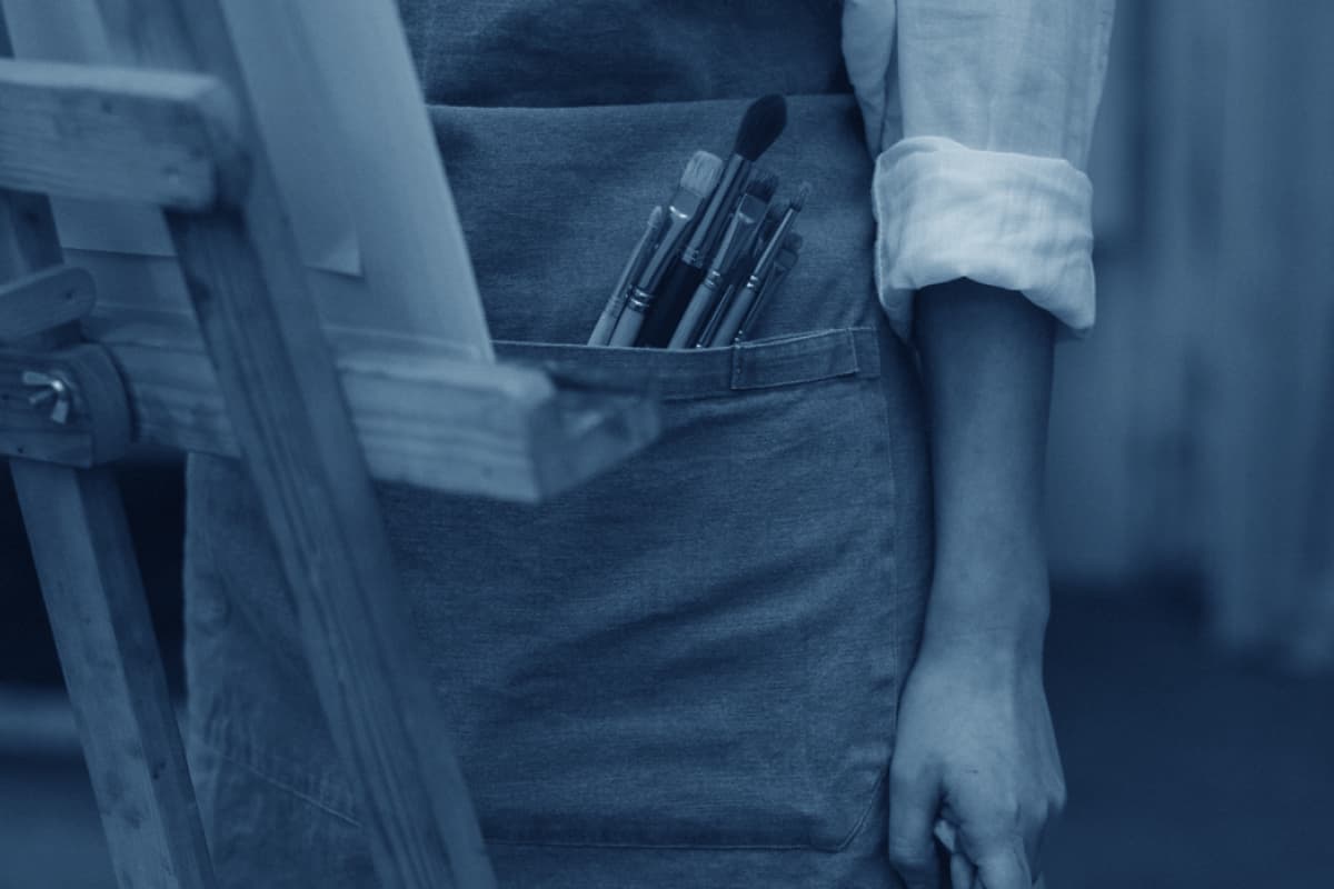 Half torso of artist in overalls with paintbrushes in her pocket and part of an easel.
