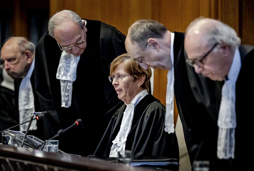 Woman in judge's attire seated, surrounded by men in judge's attire standing.
