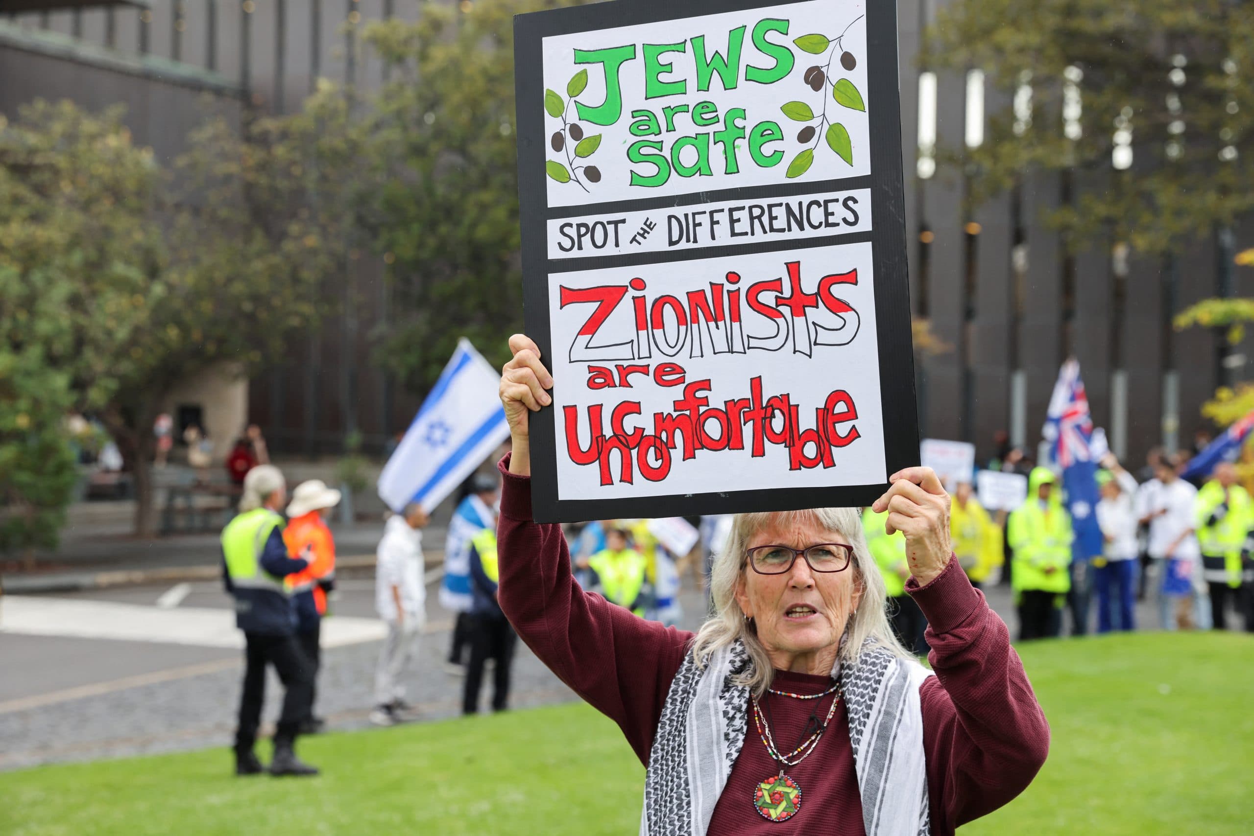 Woman wearing a keffiyeh holds up a sign reading "Jews are Safe, Zionists are uncomfortable, spot the difference".