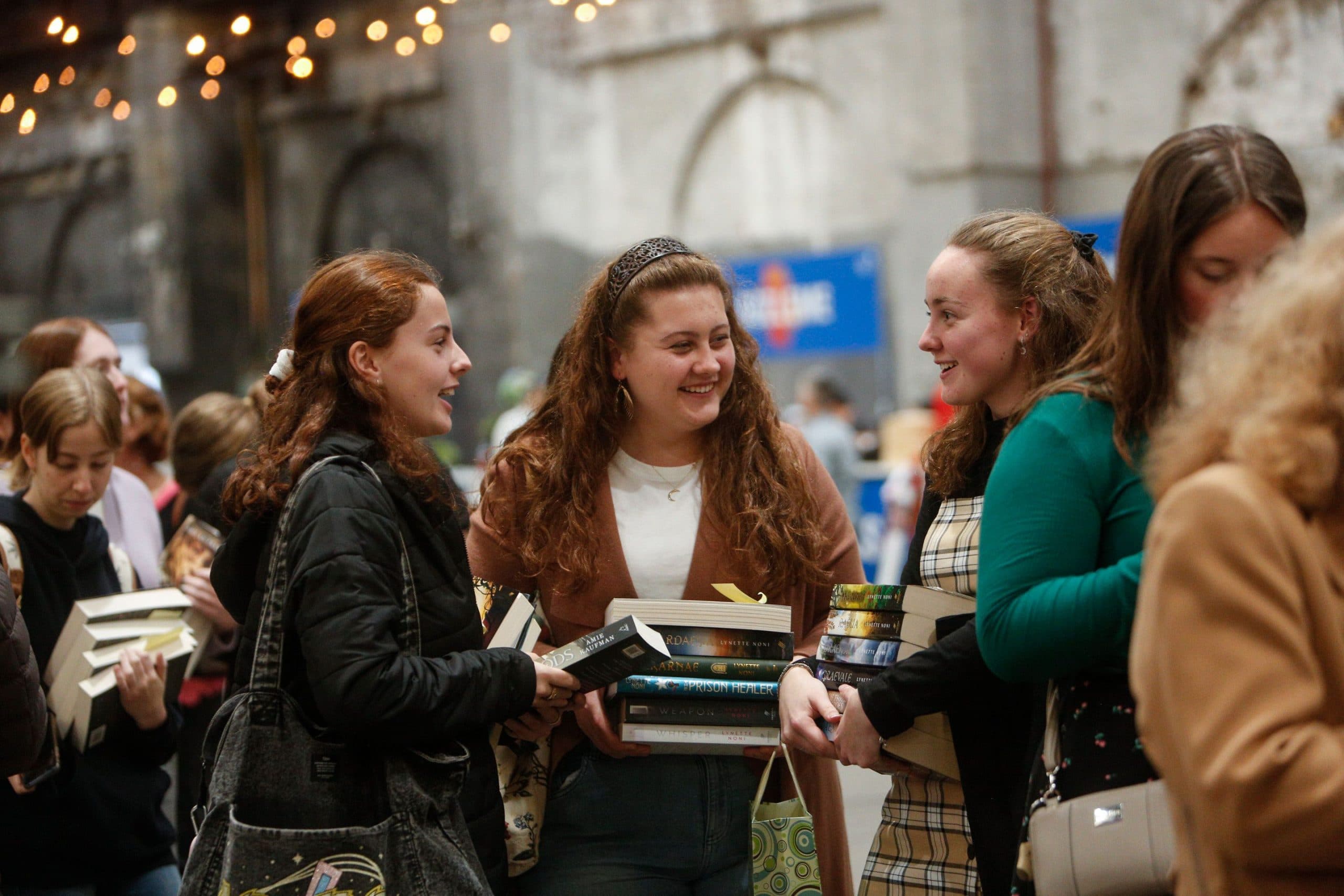 Three young women laughing and carrying piles of books