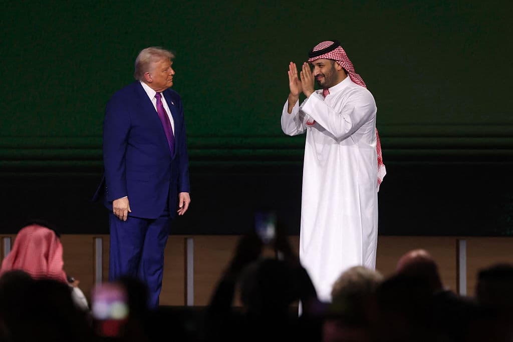 man in blue suit is clapped by man in white robe and red checked keffiyah as audience watches
