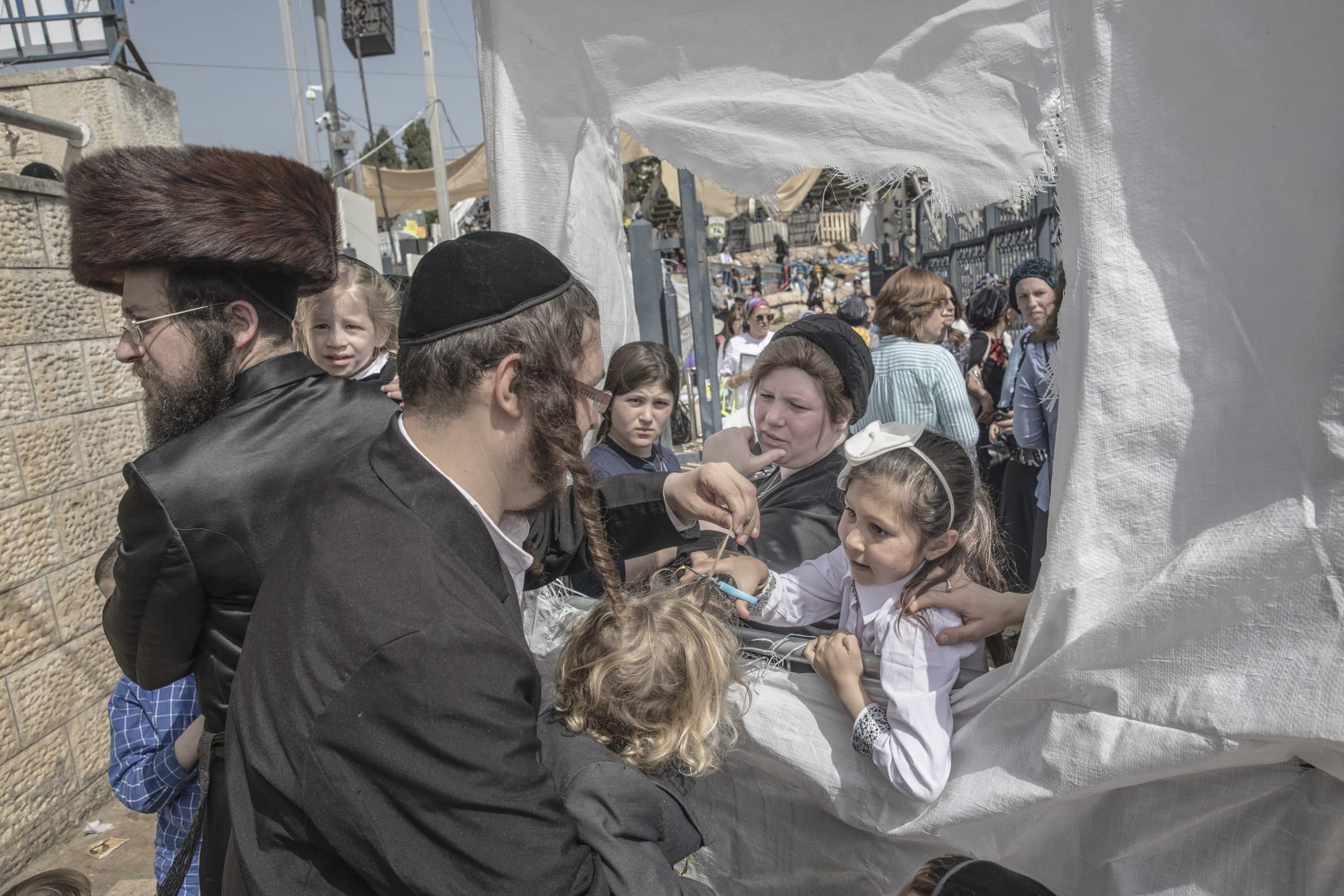 Ban with sidelocks holds small boy while girl reaches through a hole in a plastic sheet with scissors