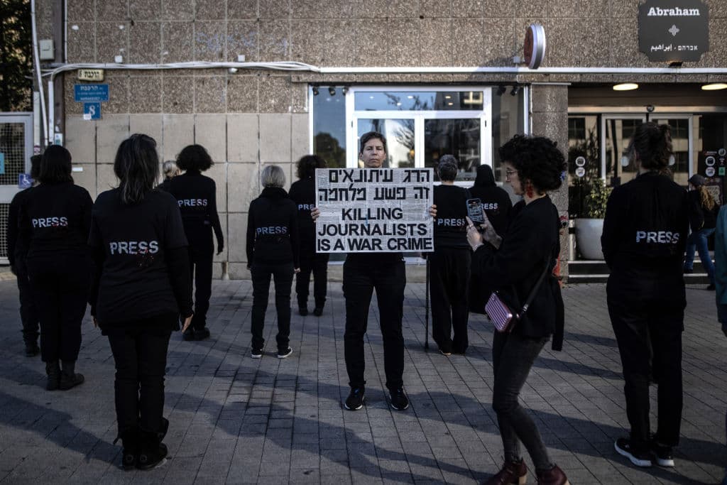 A woman in black holding a sign that reads 'Killing journalists is a war crime' in Hebrew and English, in front of several people in black with their backs to the camera and the word 'Press' printed on their shirts.
