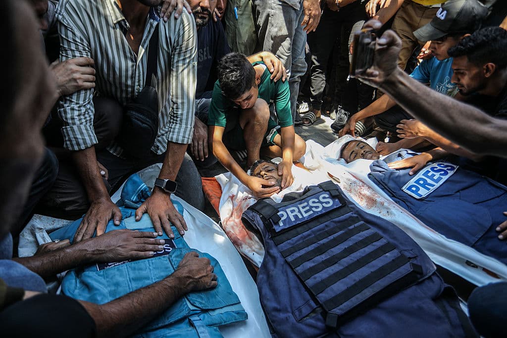 3 bodies, covered with Press vests, surrounded by mourners.