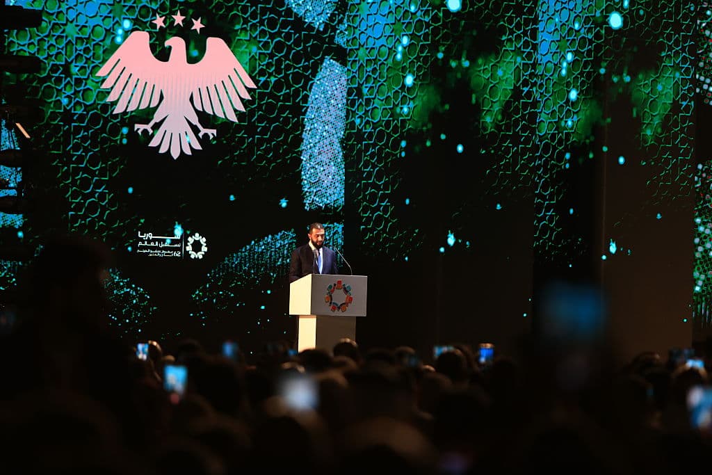 Man at a lectern in front of dramatic green and blue projections and a projection of a spread-winged eagle and 3 stars