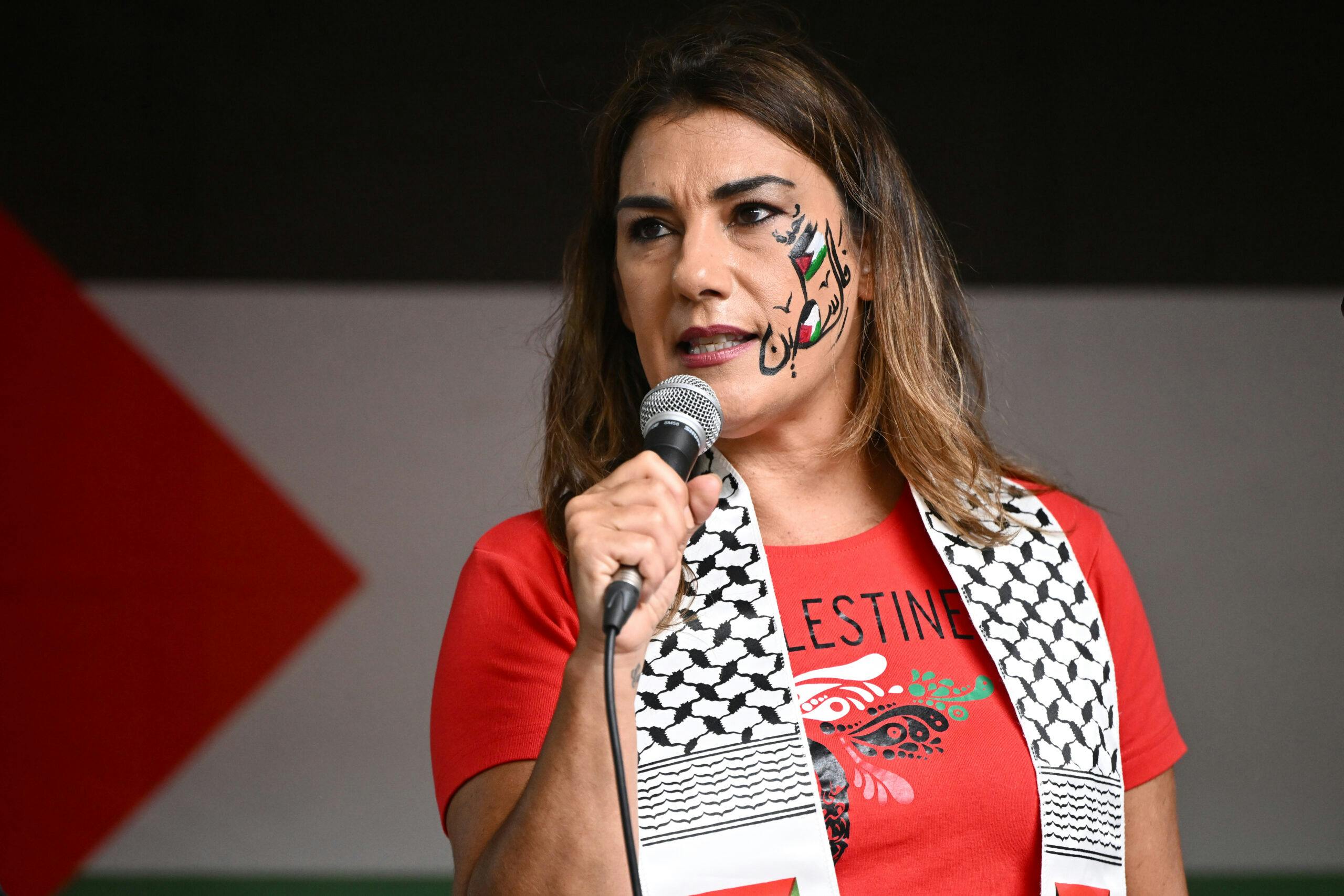 Woman holding a microphone wearing a keffiyah with a Free Palestine t-shirt and Palestine coloured face paint