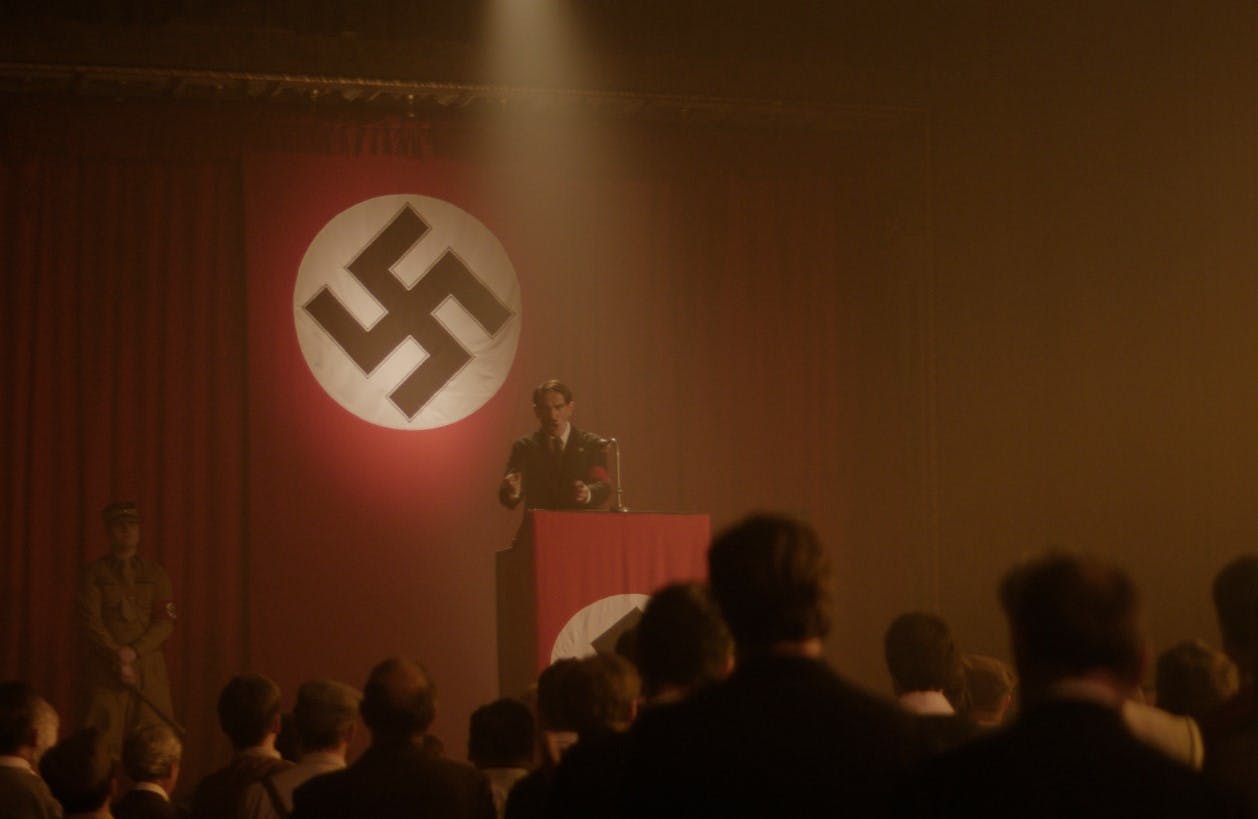 Man on podium in front of large audience with swastika flag in background