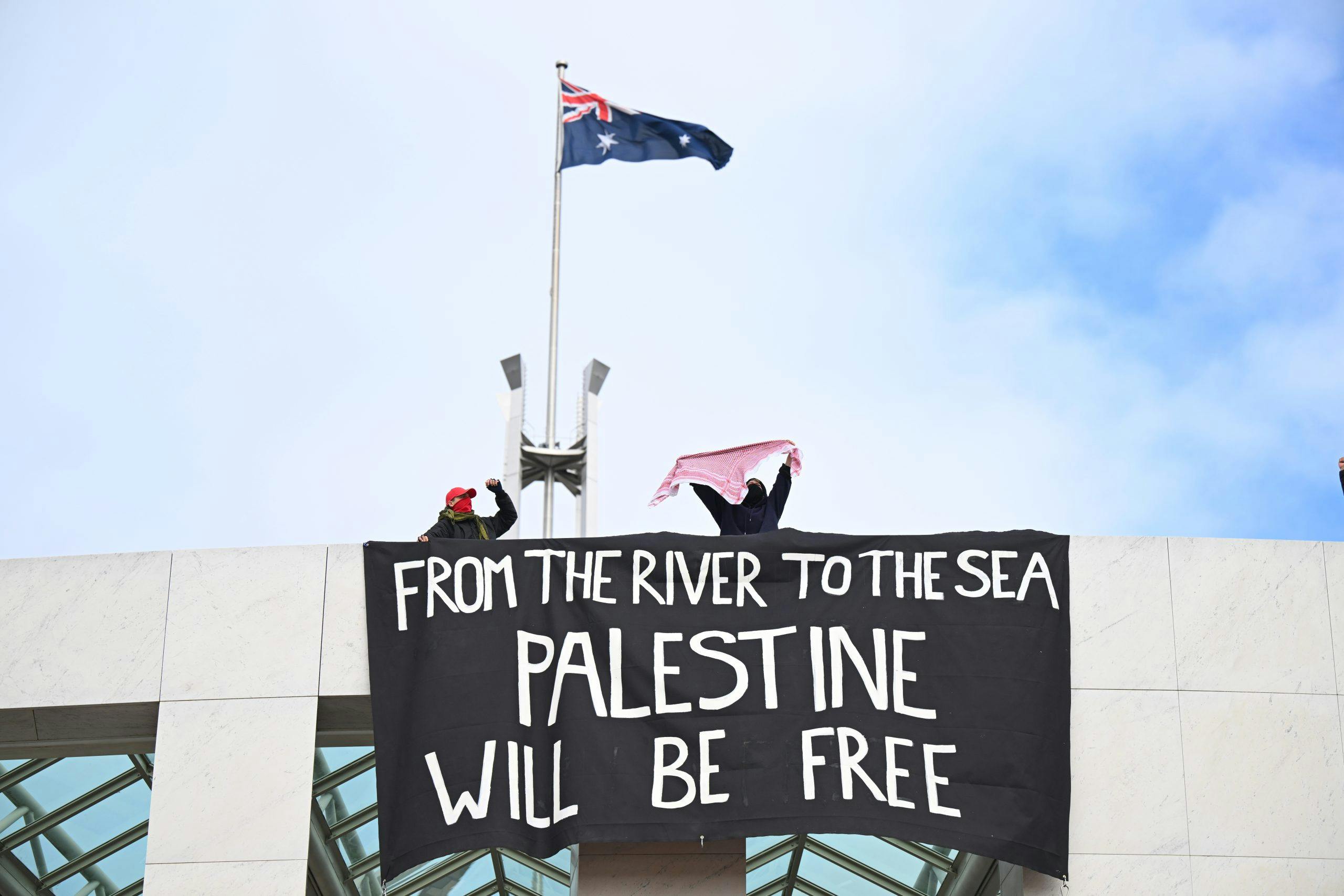 Australian flag and poster reading 'From the river to the sea Palestine will be free' displayed by people on a high wall.