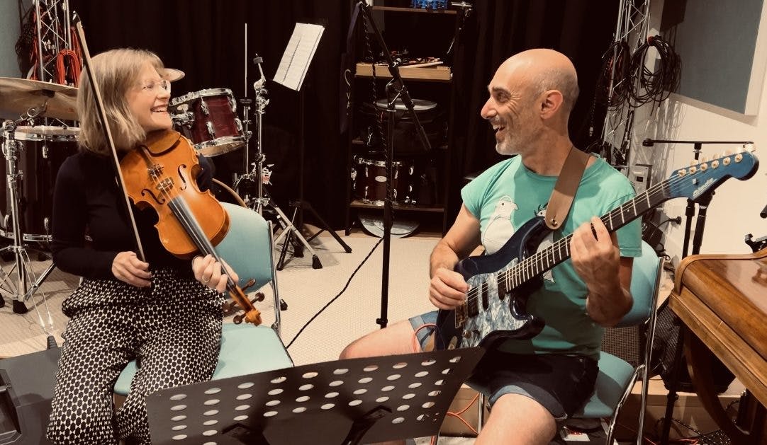 A woman smiling holding a violin and a man smiling holding a guitar in a recording studio