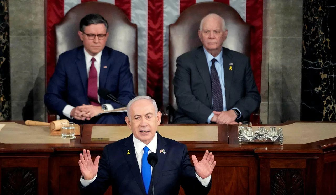 Three men, one speaking, two seated behind.