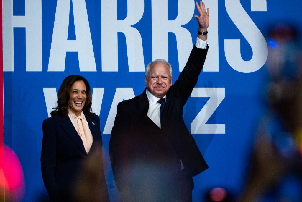 Man with raised hand and woman smiling in front of a poster reading Harris Walz