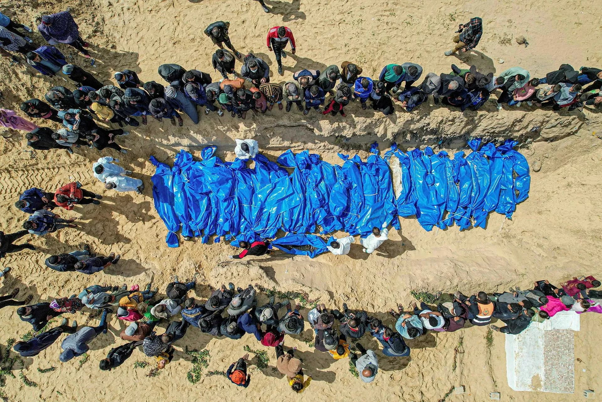 A row of body-shaped blue sacks lying on sand with people standing around.