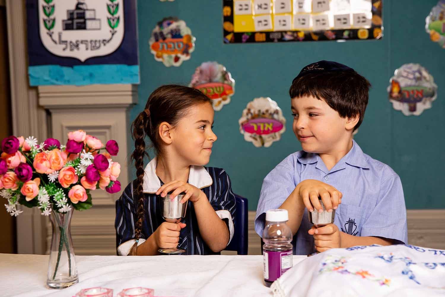 Girl and boy holding kiddush cups in front of sabbath table with children's art behind them